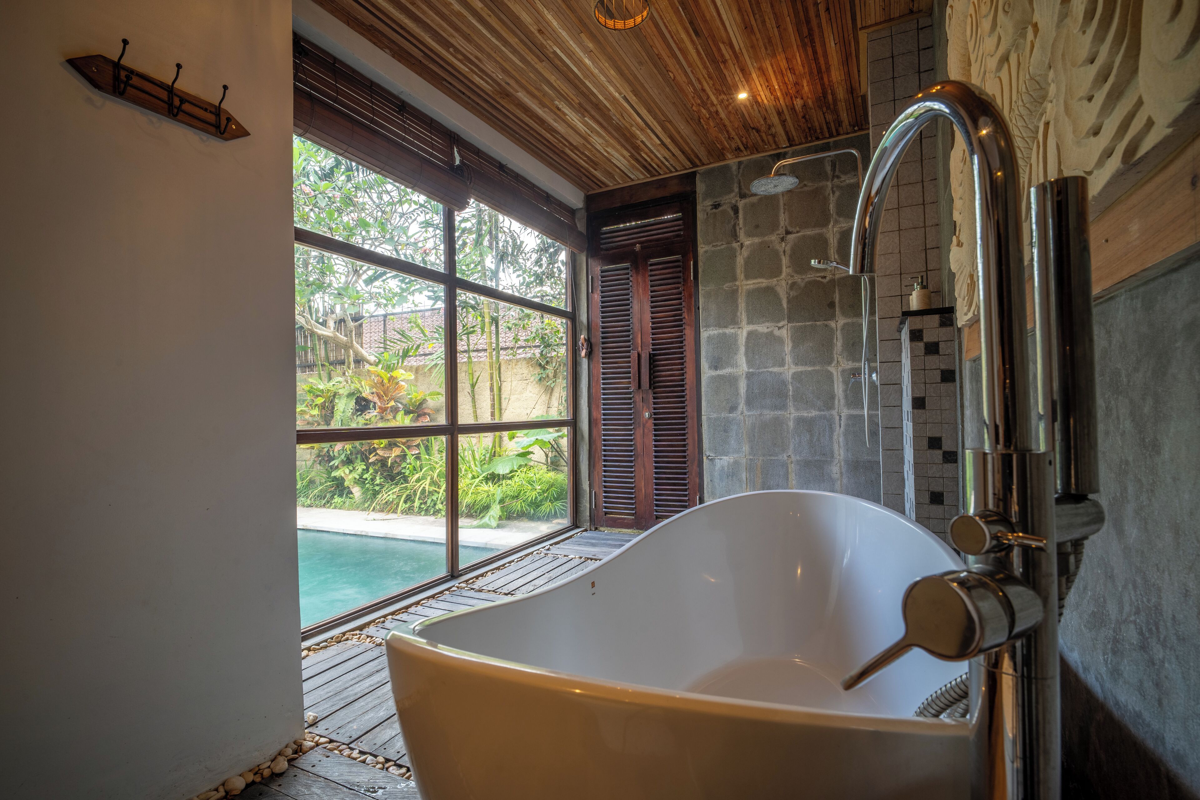 Una bañera independiente grande en un cuarto con techo de madera y vista a una piscina exterior.