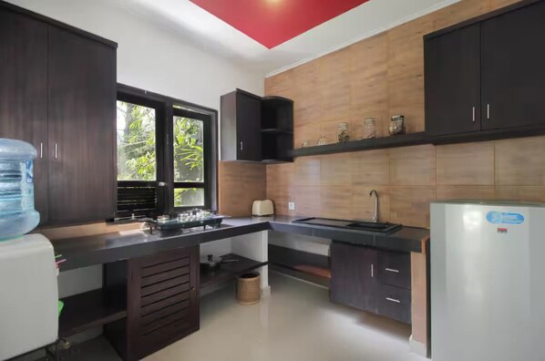 A modern kitchen with dark wooden cabinets, a black countertop, a white refrigerator, and a window with a view of greenery. - Tri Datu Villa (Ubud)