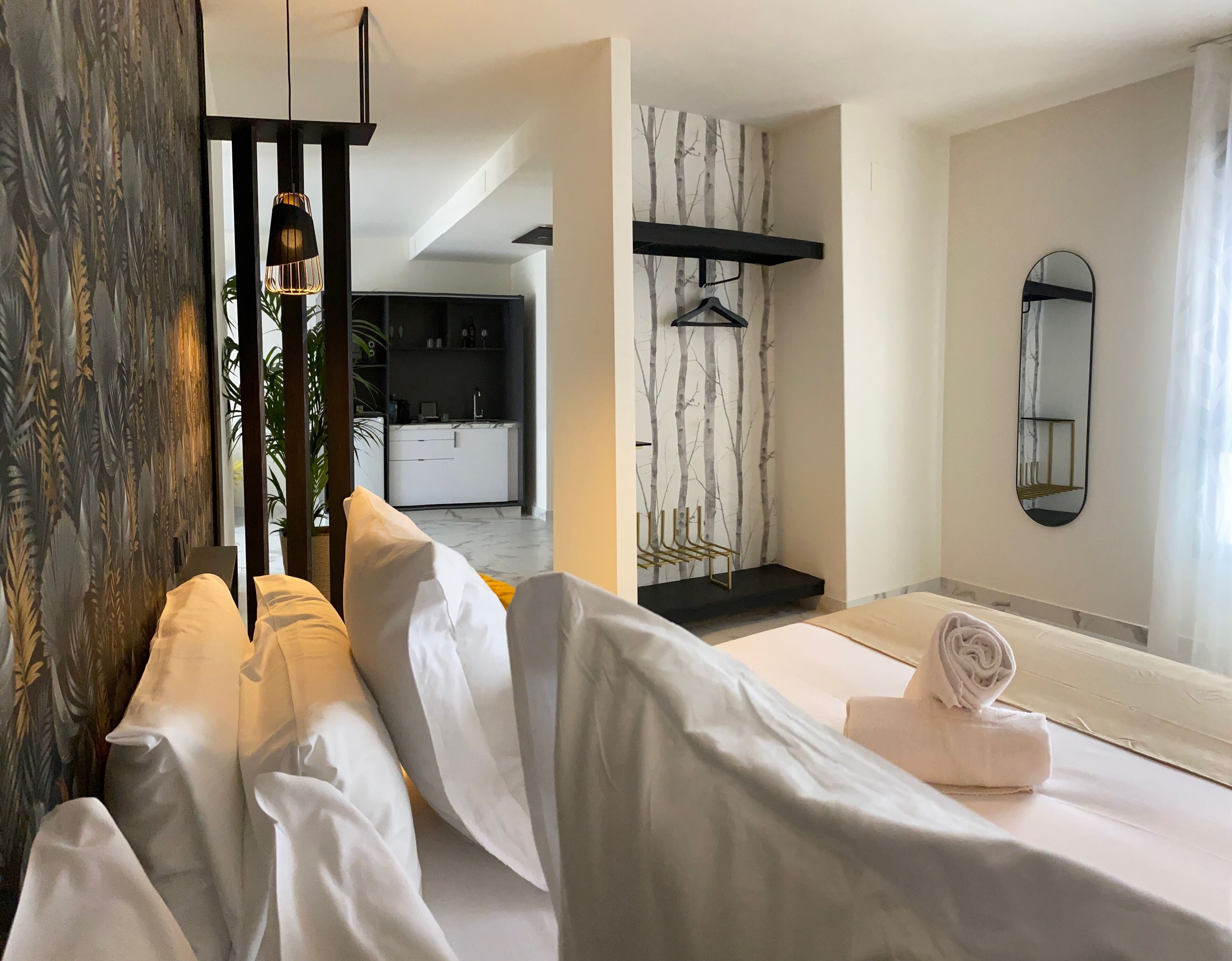 Quarto de hotel com cama, luminária pendente, espelho fixado na parede e vista para a cozinha.