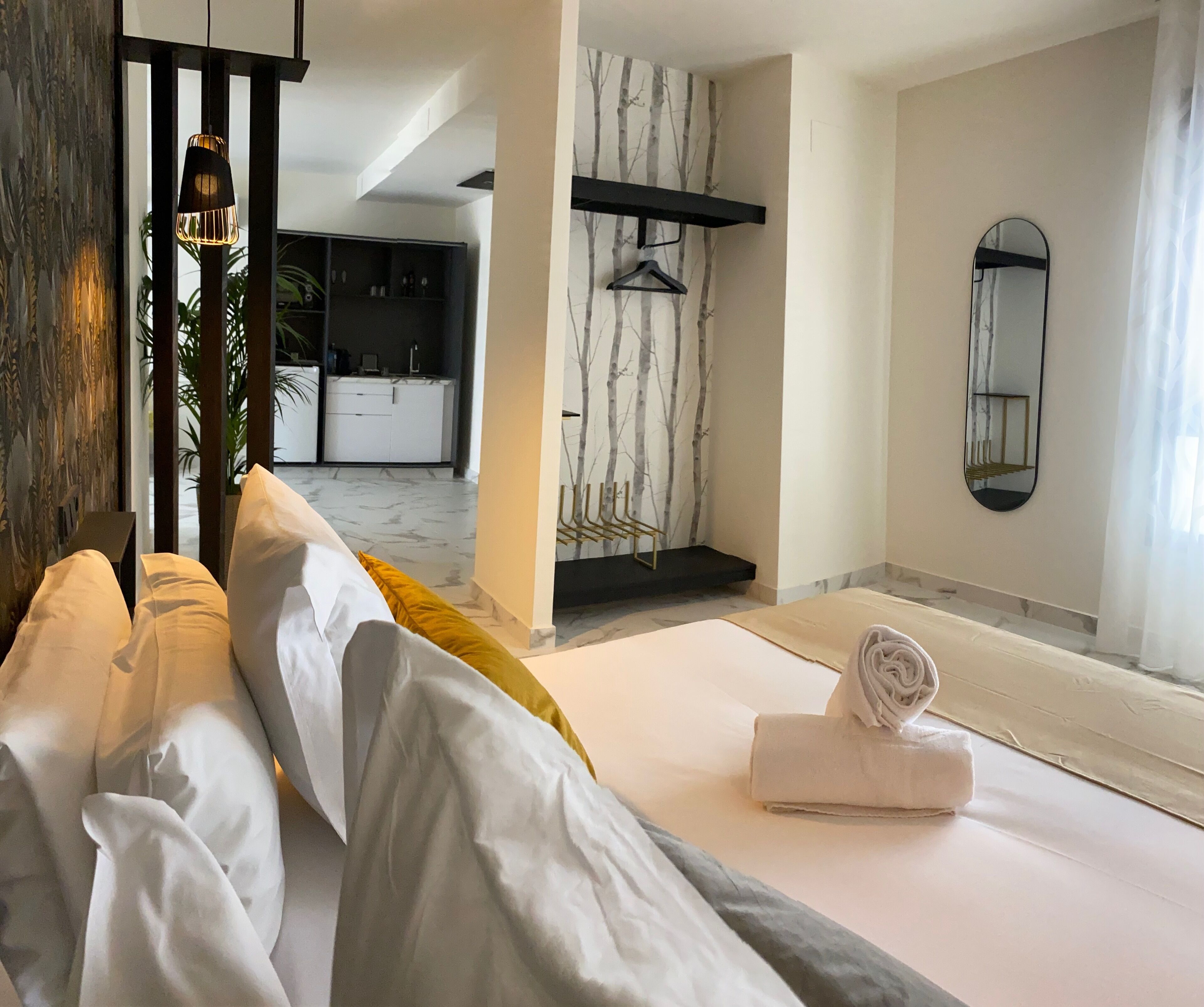 A modern hotel room with a bed, a neatly made bed with a rolled towel, a hanging light fixture, a wardrobe, a mirror, and a view of the kitchen area.