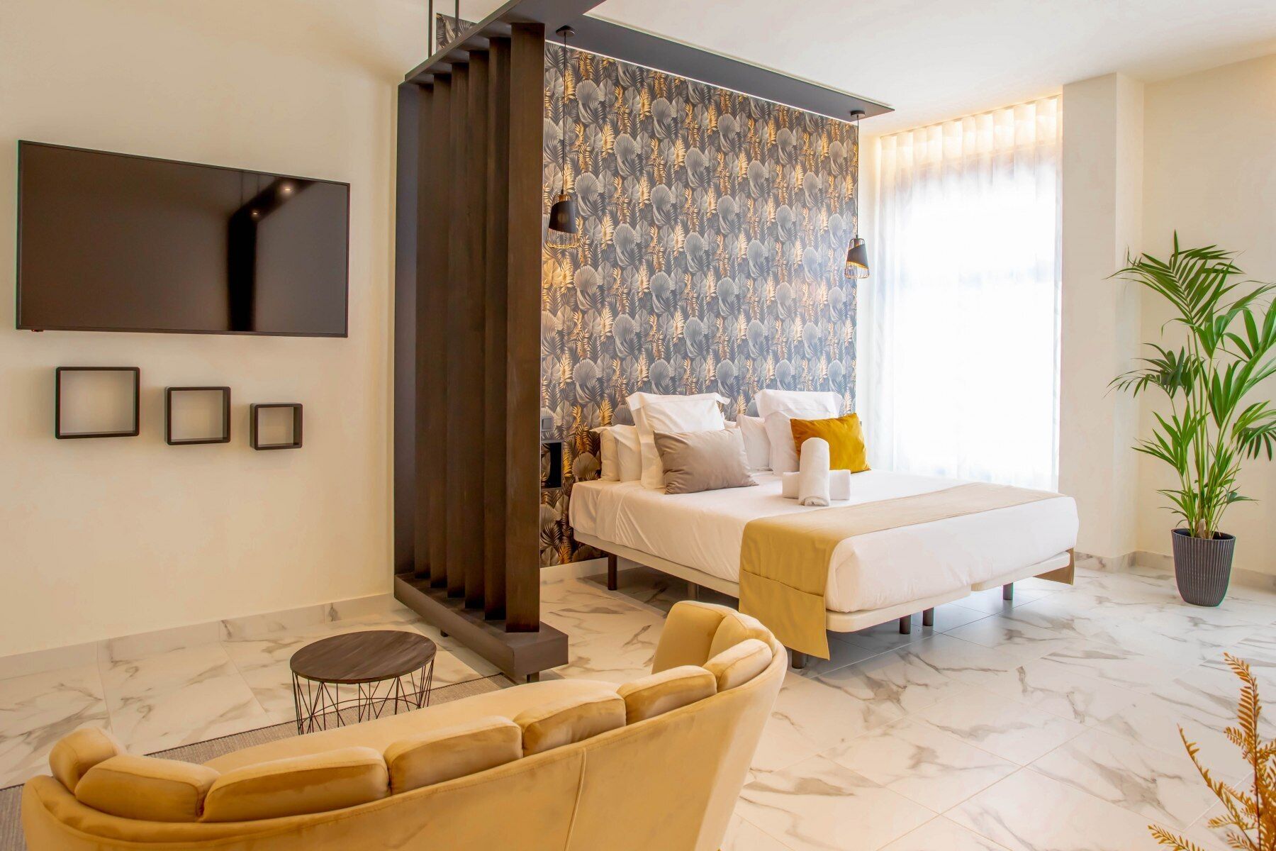 A modern hotel room with a large bed, a sofa, a small table, and a potted plant.
