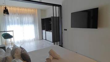 Ein modernes Schlafzimmer mit Bett, einem an der Wand montierten Fernseher und Blick in den Wohnbereich.