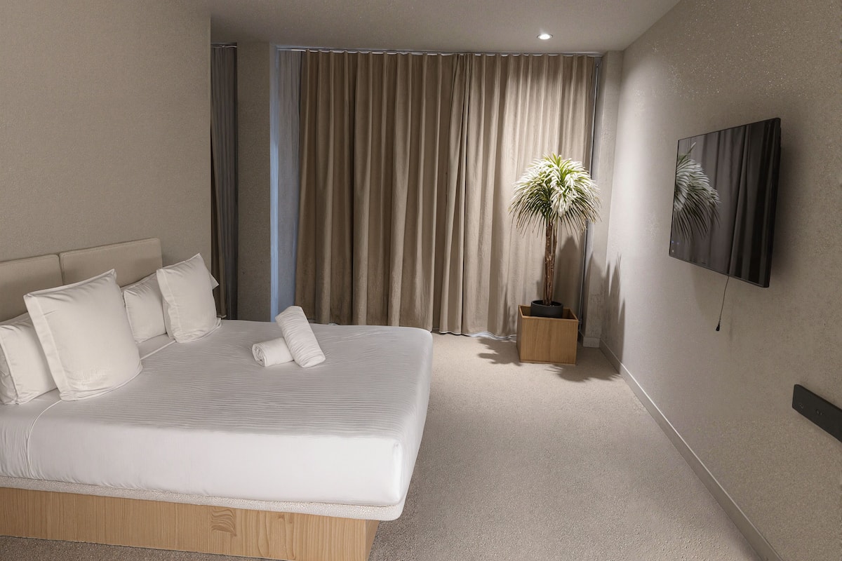 Une chambre d’hôtel moderne dotée d’un grand lit, d’une plante en pot, d’un téléviseur à écran plat et d’une grande fenêtre avec des rideaux.
