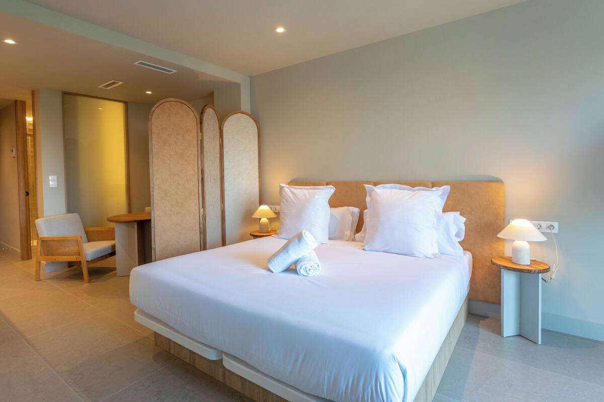 Un lit bien fait, avec du linge de lit blanc et une serviette enroulée, deux lampes de chevet et une chaise en bois, dans une chambre d’hôtel moderne.