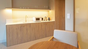 Une cuisine moderne avec un plan de travail blanc, un four à micro-ondes, une machine à café et un évier. On trouve un meuble en bois au-dessus du plan de travail et une chaise devant.