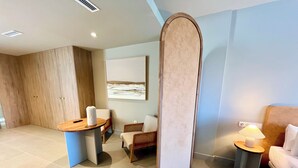 Une chambre d’hôtel moderne avec une armoire en bois, une table ronde dotée d’une télécommande, deux chaises, un lit avec une tête de lit, une petite table de chevet avec une lampe et un grand tableau abstrait accroché au mur.