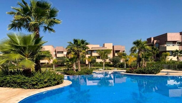 Una piscina de borde curvo, rodeada de palmeras y un edificio con varios balcones.