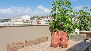 Una terraza en la azotea con mesa y sillas de madera, macetas de terracota y vistas de la ciudad.