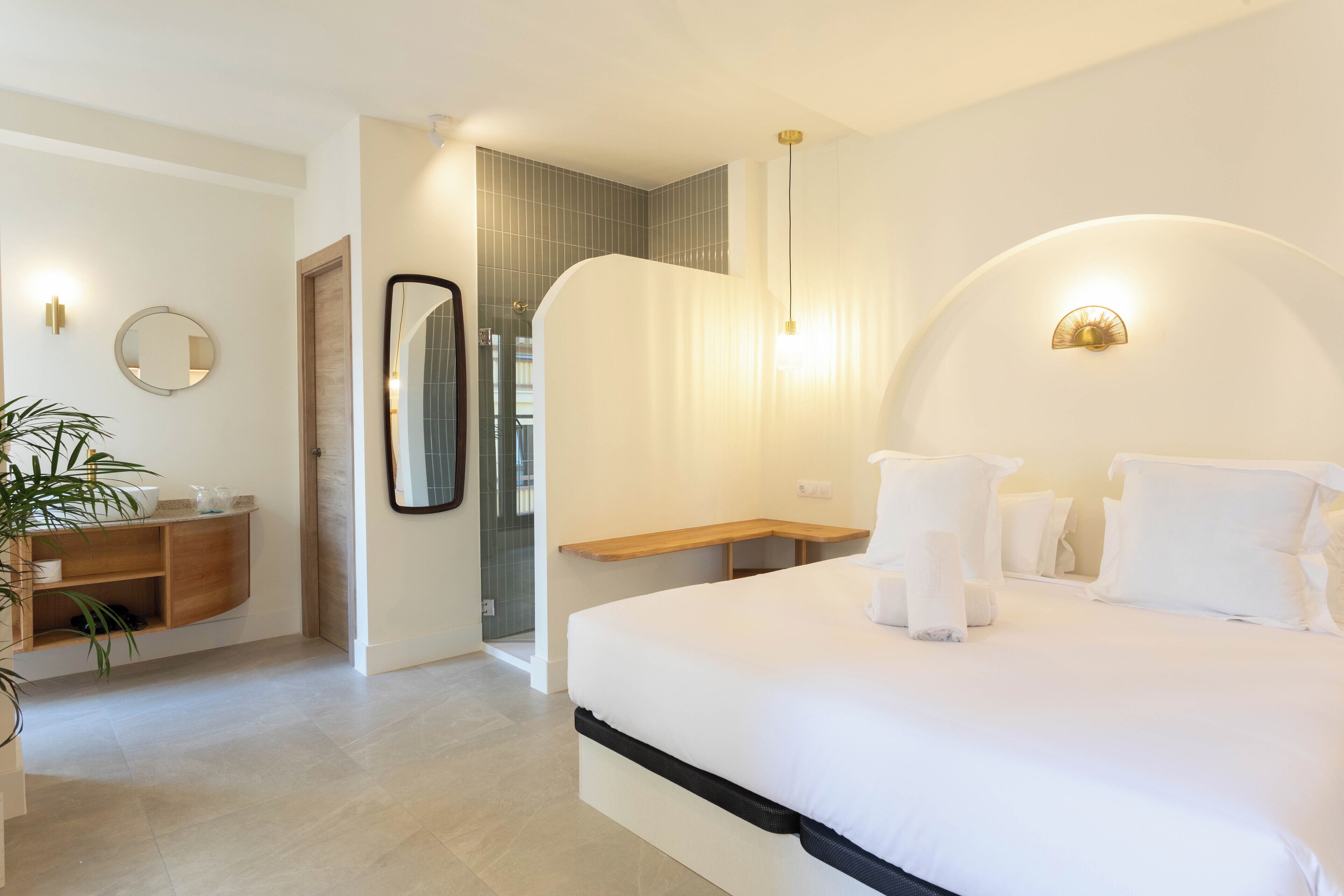 Una habitación de hotel moderna con una cama grande, una mesita de noche, un espejo, un banco de madera y una planta en maceta.