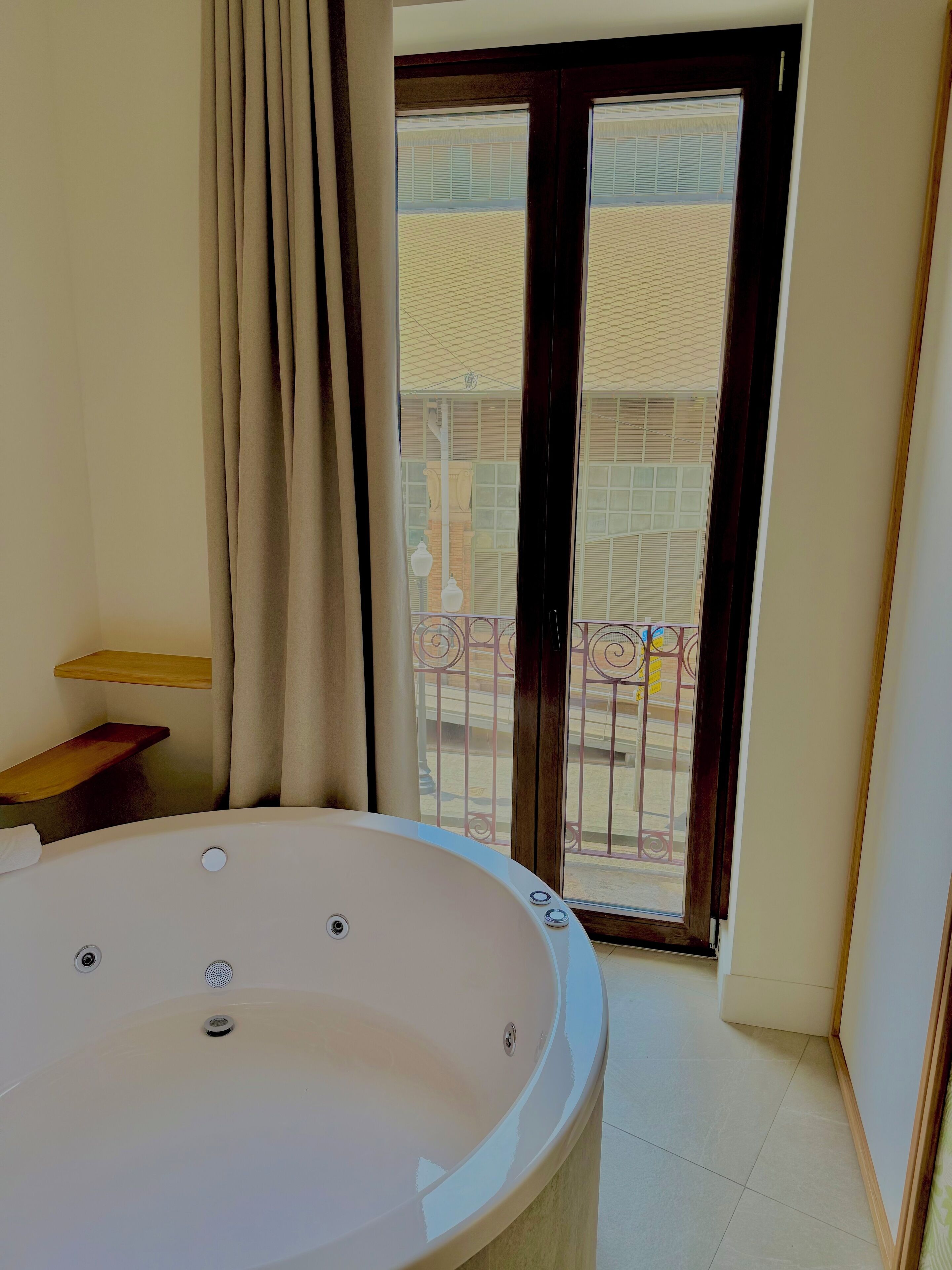 Bany amb banyera gran, dutxa i vàter. Hi ha una finestra amb cortines i vista a un balcó amb barana.