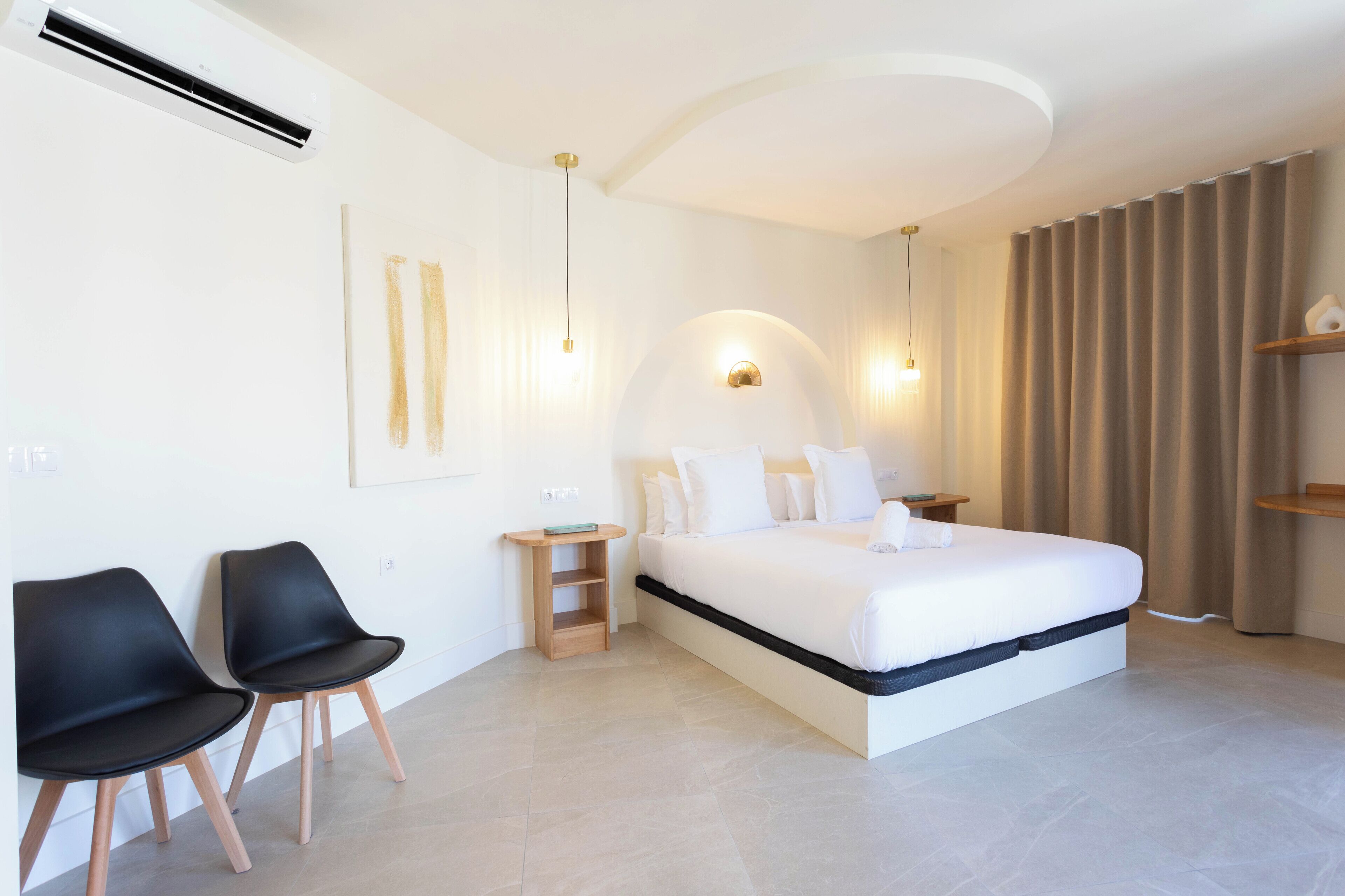 Ein modernes Schlafzimmer mit einem großen Bett, zwei schwarzen Stühlen, einem Nachttisch und einer Wand-Klimaanlage.