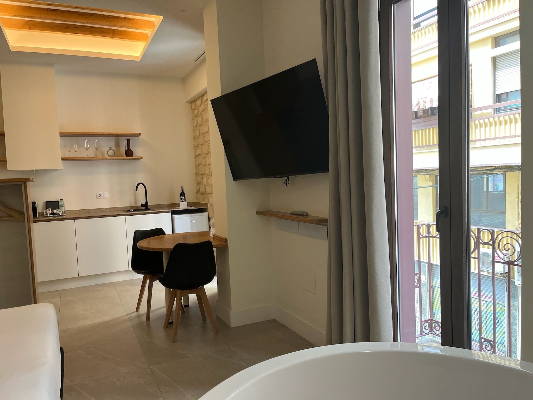 A modern kitchen with white cabinets, a dining area with a table and chairs, a flat-screen TV mounted on the wall, and a balcony with metal railings.