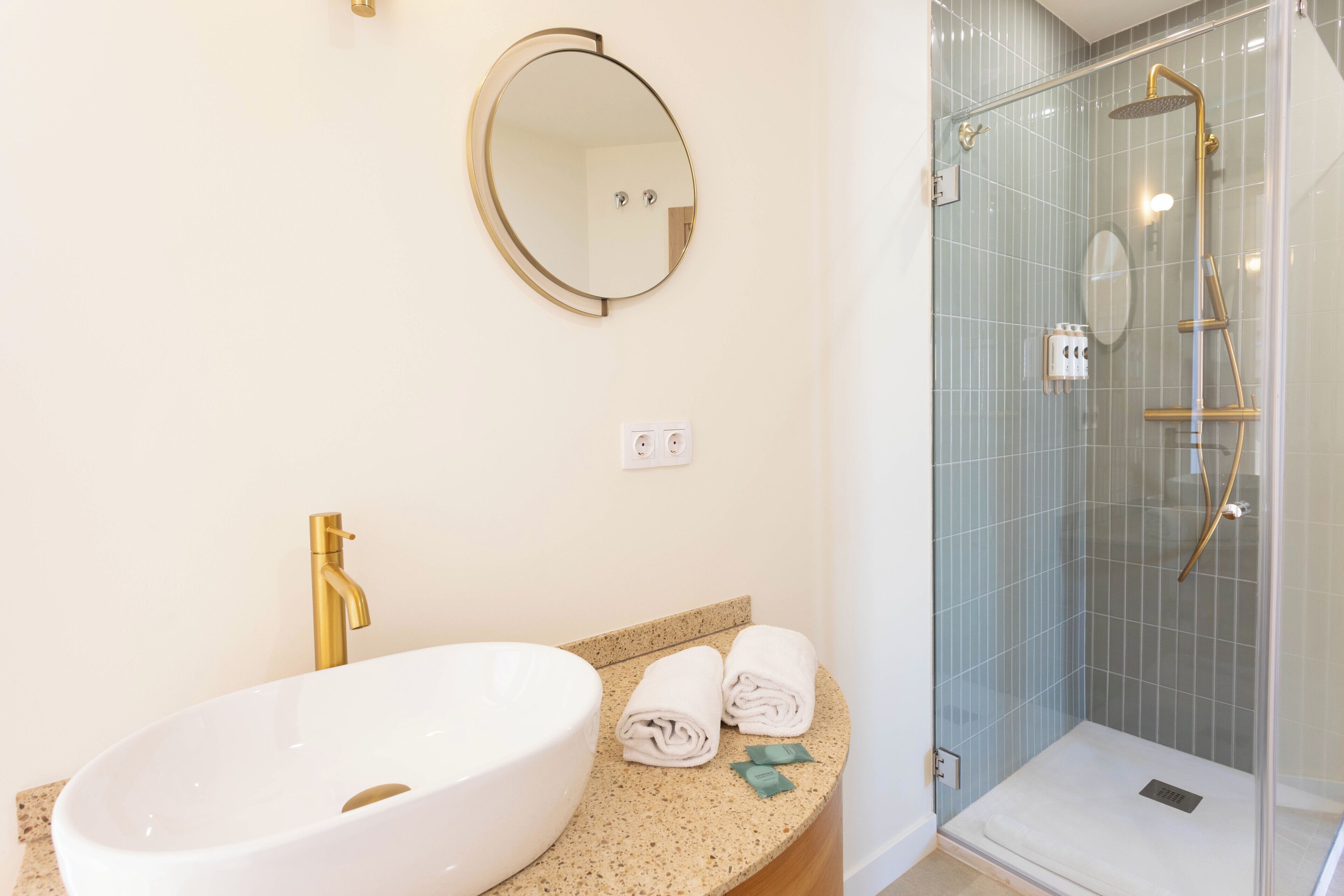 A bathroom with a white sink, gold faucet, and a round mirror. There are towels on the counter and a glass-enclosed shower with gold fixtures.