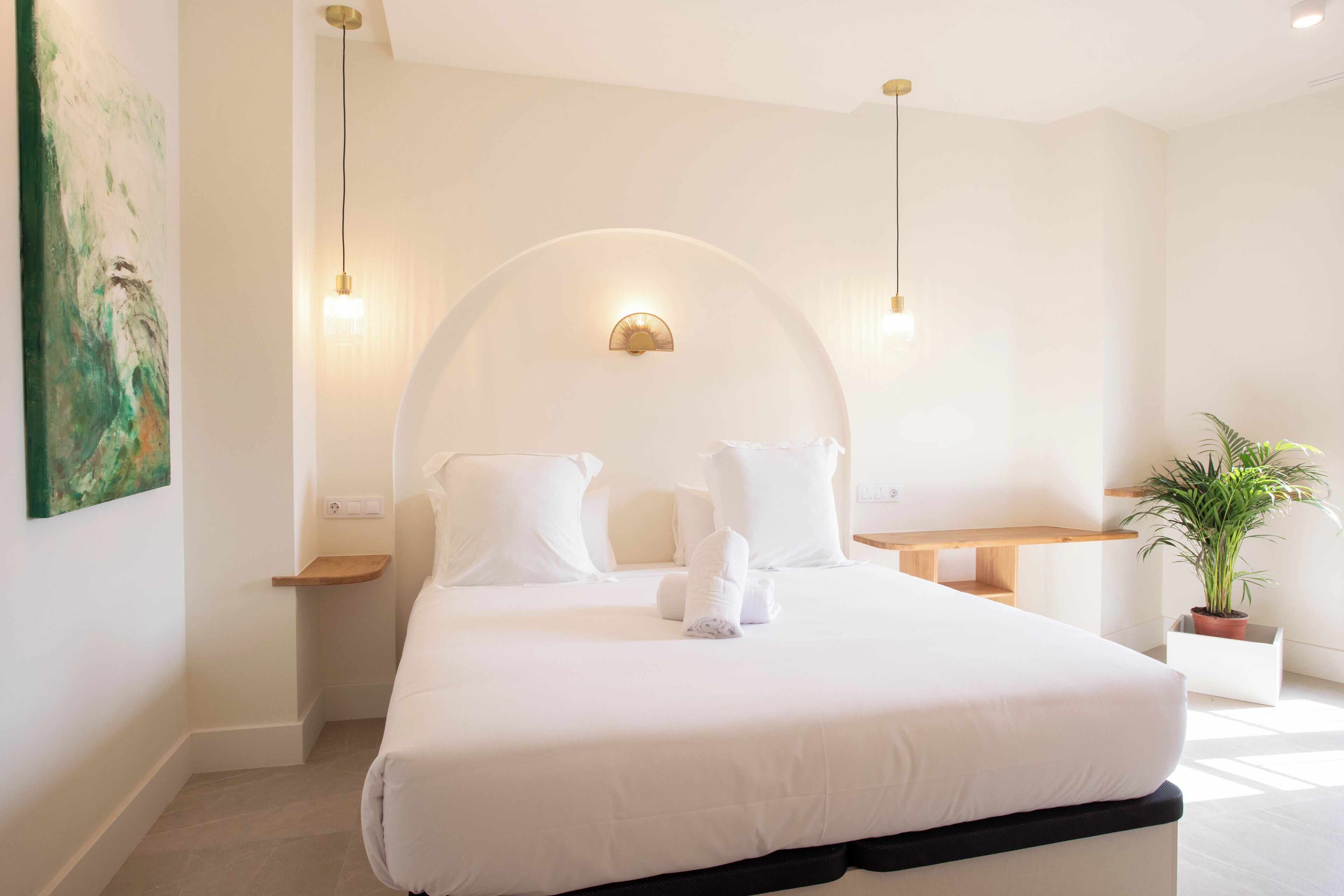 A modern hotel room with a large bed, two bedside tables, a potted plant, and a wall-mounted light fixture.