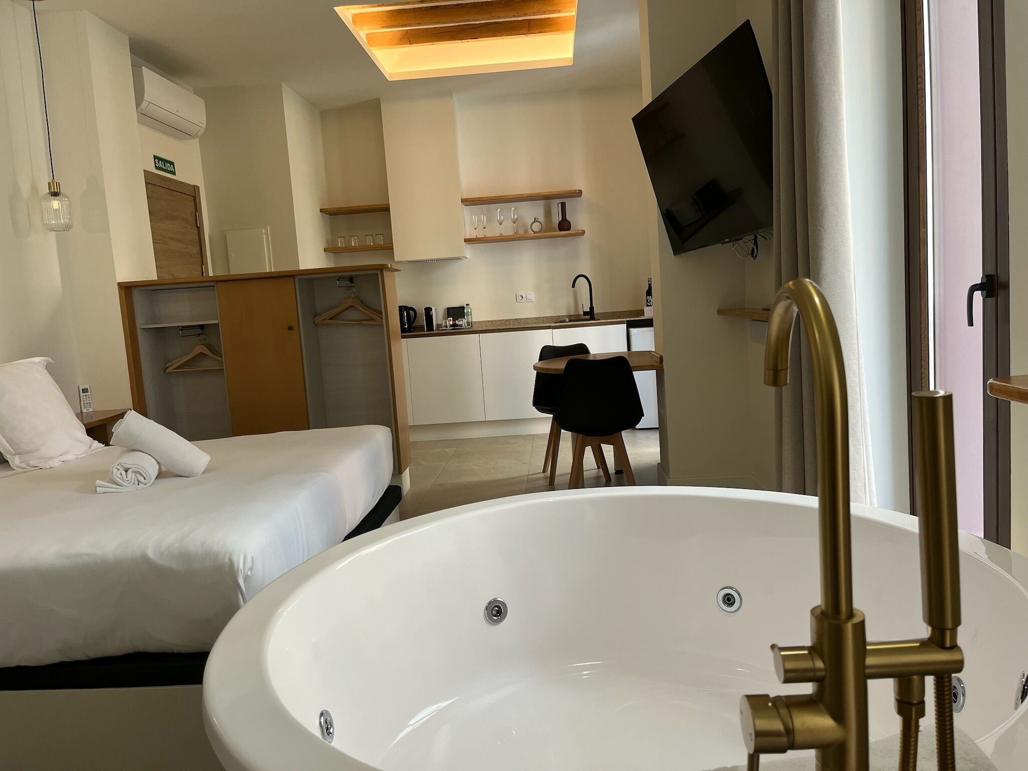 A hotel room with a large bathtub, a bed, a desk with a chair, a TV mounted on the wall, and a kitchenette area.