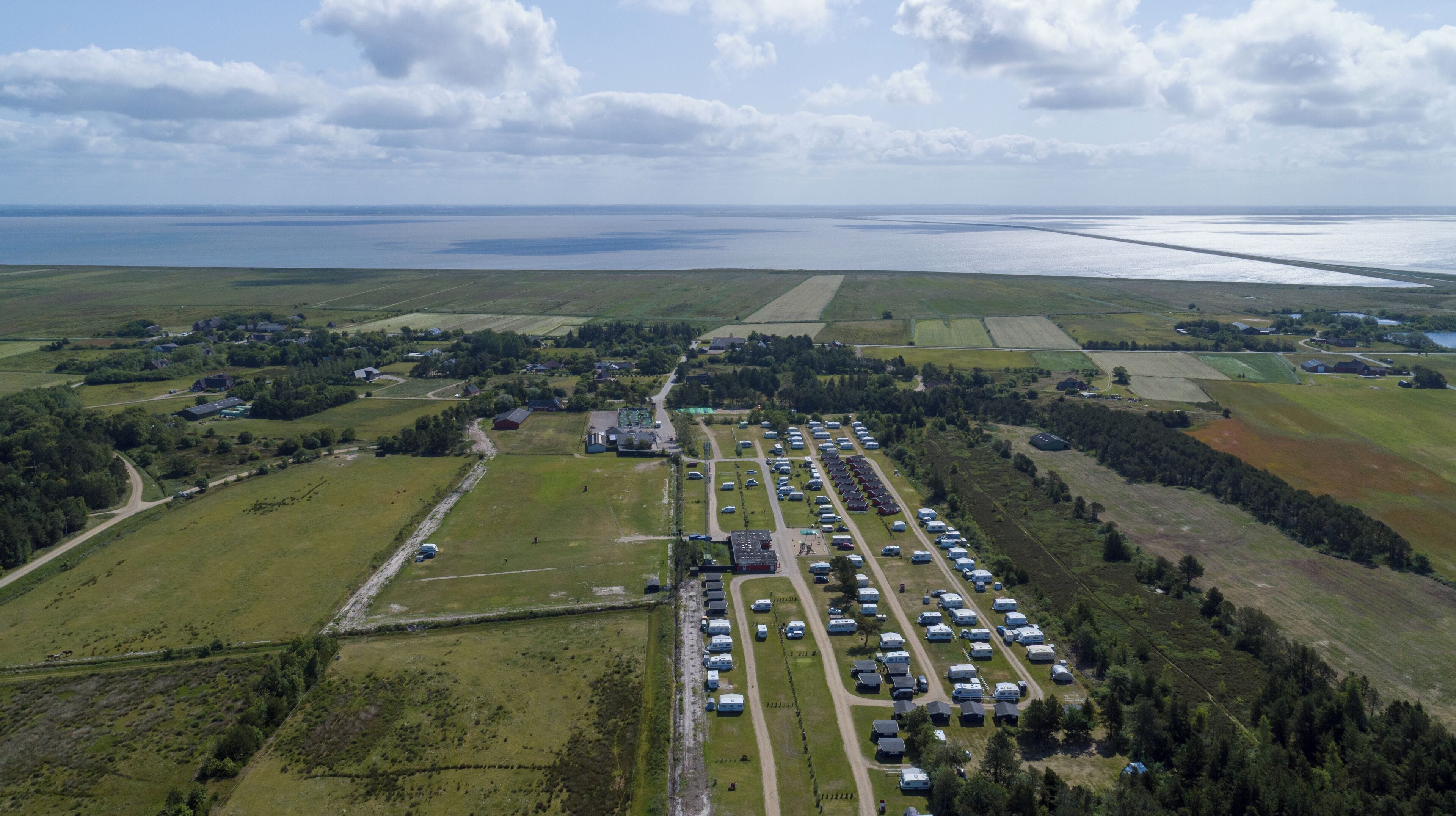 Ett flygbildsbild av en campingplats med husbilar parkerade på rader, omgiven av åkrar och en sjö eller ett annat vattendrag i bakgrunden.