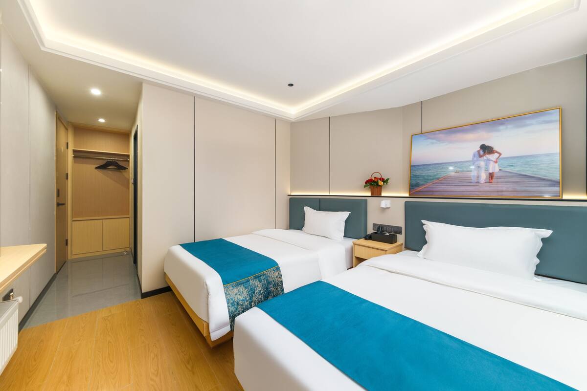 A hotel room with two beds, a wooden floor, a wardrobe, and a painting of a couple on a pier.