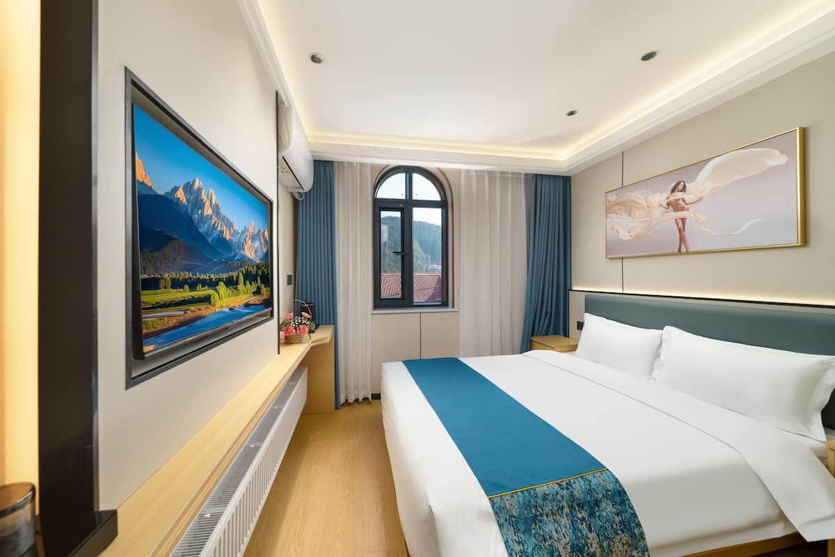 A hotel room with a large bed, a TV mounted on the wall, a window with curtains, and a painting above the bed.