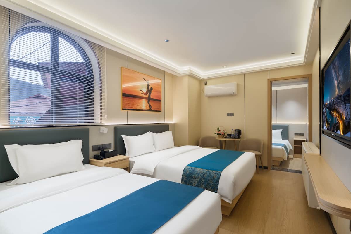 A hotel room with two beds, a TV, a desk, and a window with blinds.