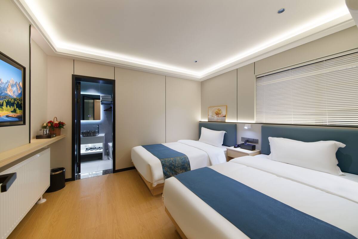 A hotel room with two beds, a desk, a mirror, and a window with blinds.