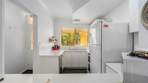 A compact kitchen with white cabinets, a sink, a window with blinds, and a refrigerator. - Shell Cove by Charlesworth (Korora)