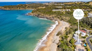 An aerial view of a coastal area with a beach, houses, and a sign with a palm tree symbol. - Shell Cove by Charlesworth (Korora)