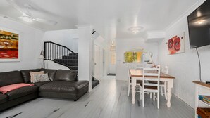 A modern living room with a black leather sofa, a dining table with white chairs, a black spiral staircase, and a flat-screen TV mounted on the wall. - Shell Cove by Charlesworth (Korora)