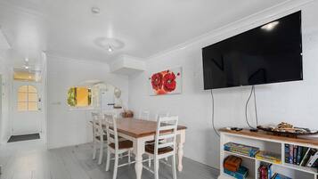 A dining area with a wooden table and white chairs, a flat-screen TV mounted on the wall, and a bookshelf with board games.