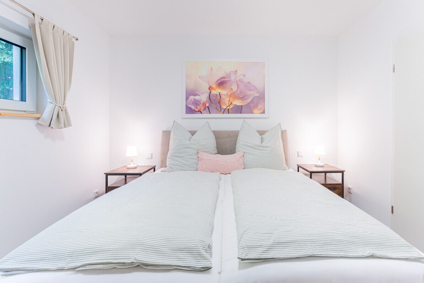 Ein Doppelbett mit gestreifter Bettwäsche, zwei Nachttische mit Lampen, ein Fenster mit Vorhängen und ein Blumenbild an der Wand.