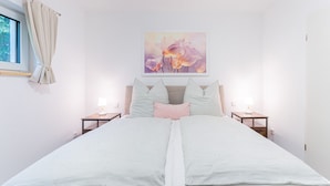 A double bed with striped bedding, two bedside tables with lamps, a window with curtains, and a floral painting on the wall. - Sund Huus Altefähr - Urlaub im Süden der Insel (Altefähr)