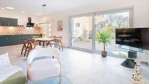 A modern kitchen with dark green cabinets, a dining area with wooden chairs and a glass table, and a large sliding glass door leading to a patio with a potted plant and a dog statue. - Sund Huus Altefähr - Urlaub im Süden der Insel (Altefähr)