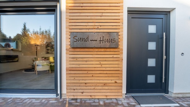 A building facade with a wooden panel wall, a sign reading "Sund-Haus," and a black door with frosted glass panels. - Sund Huus Altefähr - Urlaub im Süden der Insel (Altefähr)