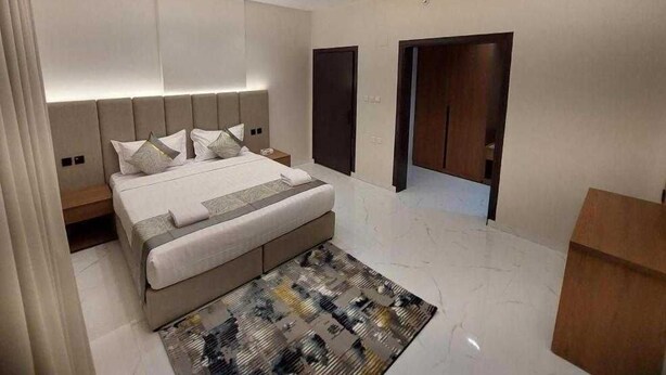 A modern bedroom with a large bed, a headboard, a bedside table, and a nightstand. There are two doors and a wooden cabinet. - Palette Luxury Doolv Serviced Apartments Tabuk (Tabuk)