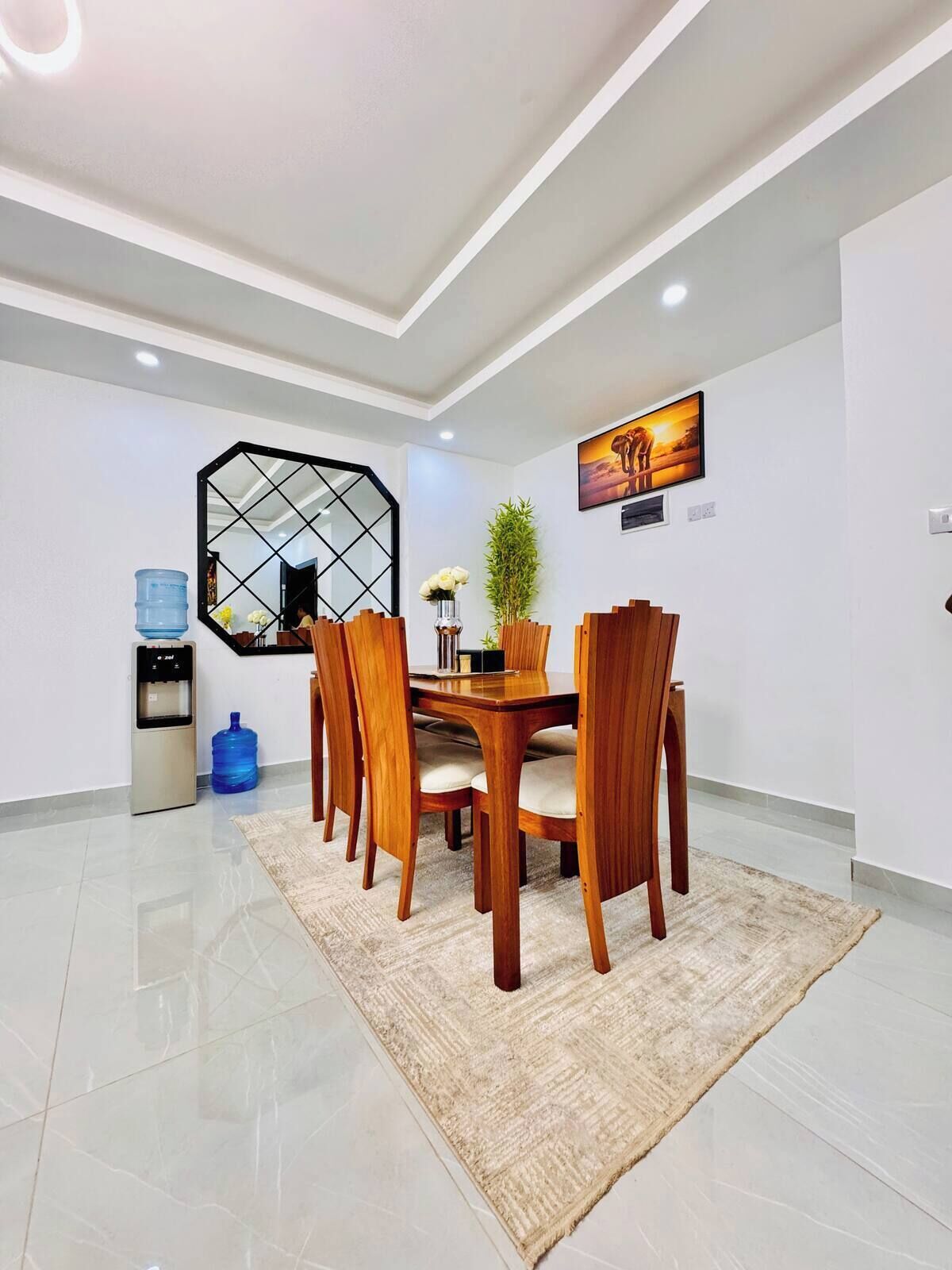 Un espace repas moderne avec une table ronde en bois, six chaises, un miroir hexagonal, un distributeur d’eau et une grande bouteille d’eau.