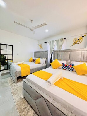 A hotel room with two beds, a ceiling fan, a large window with curtains, and a decorative wall piece. - Modern 3BR Nyali Stay, AC, Kids’ Play Area, Pool, Gym & 3 Min to Beach (Mombasa)