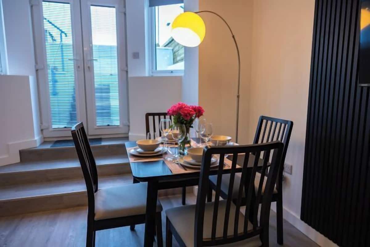 Un comedor con una mesa dispuesta para cuatro, sillas, un florero con flores y una lámpara de pie.