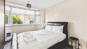 Ein Hotelzimmer mit einem Bett, einem Fenster mit Jalousien, einem Wandventilator und einem kleinen Tisch mit Stuhl.