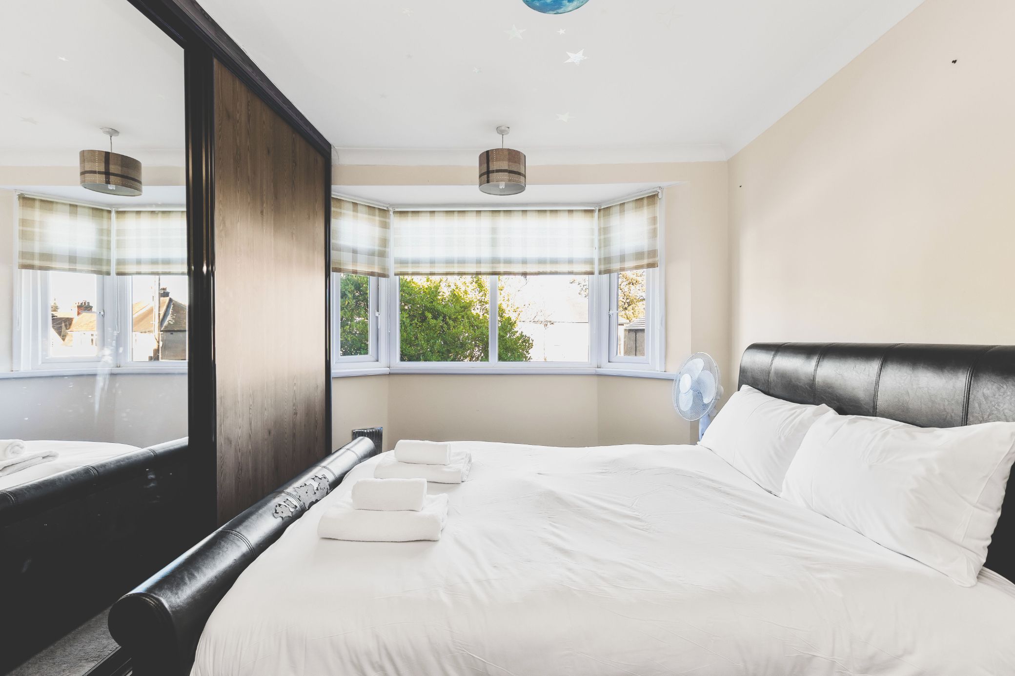 A hotel room with a large bed, a mirror, a window with a view of greenery, and a ceiling fan.