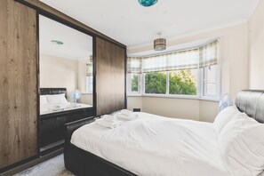 Un dormitorio con un espejo grande, una cama con ropa de cama blanca, una mesita de noche y una ventana con cortinas.