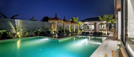 A swimming pool with lounge chairs and umbrellas, surrounded by a landscaped garden and a building with large windows.