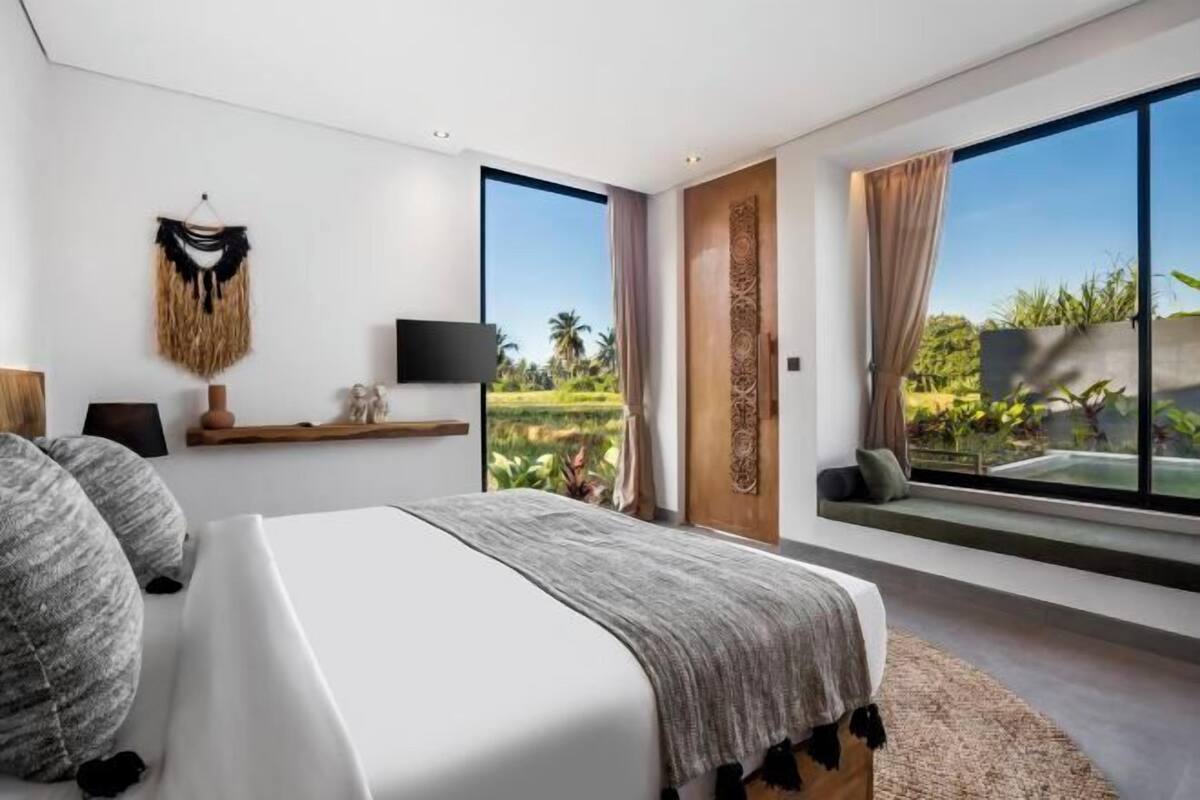 A modern bedroom with a large bed, a TV, a window with a view of greenery, and a decorative wall piece.