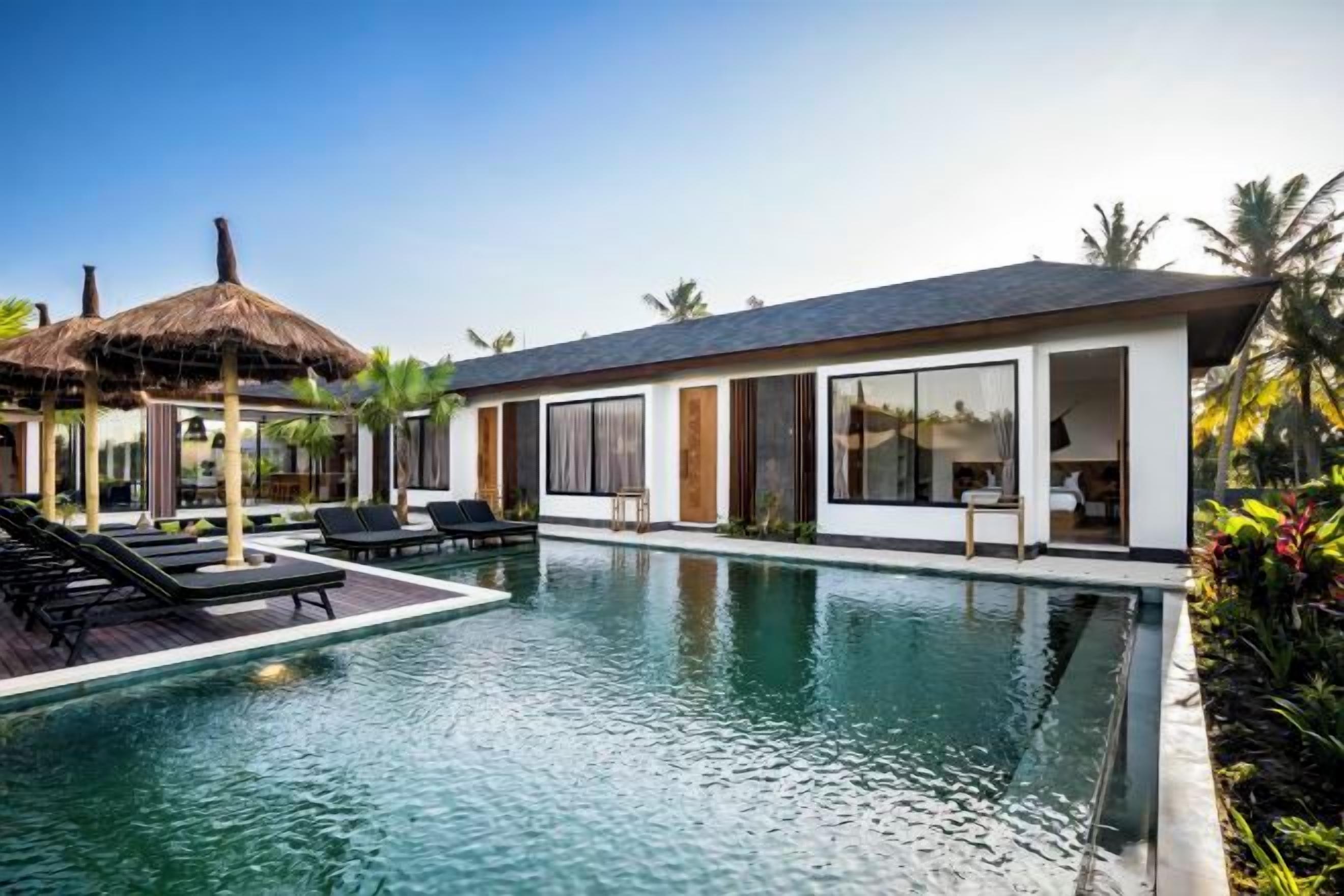 A modern resort with a swimming pool, lounge chairs, and a thatched-roof structure.