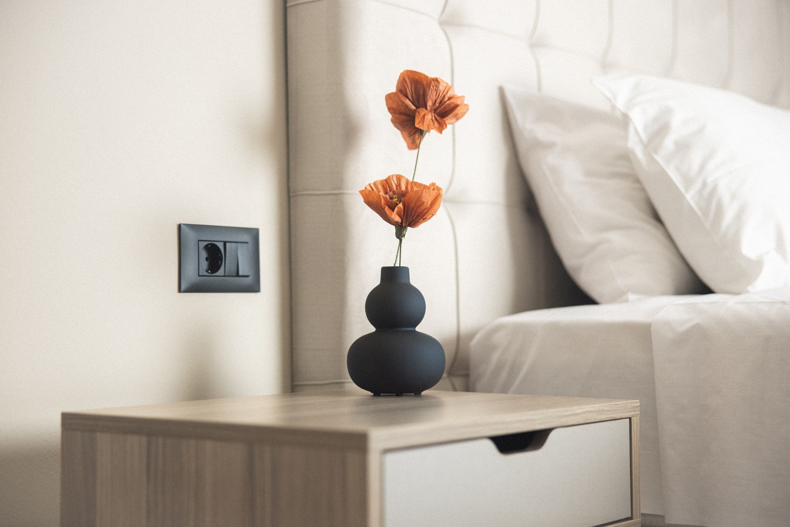 A bedside table with a vase of flowers and a framed picture on a wooden nightstand.