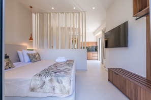 A modern bedroom with a large bed, a TV, and a view to the kitchen. - Manthos Beachfront Apartments (Laganas)