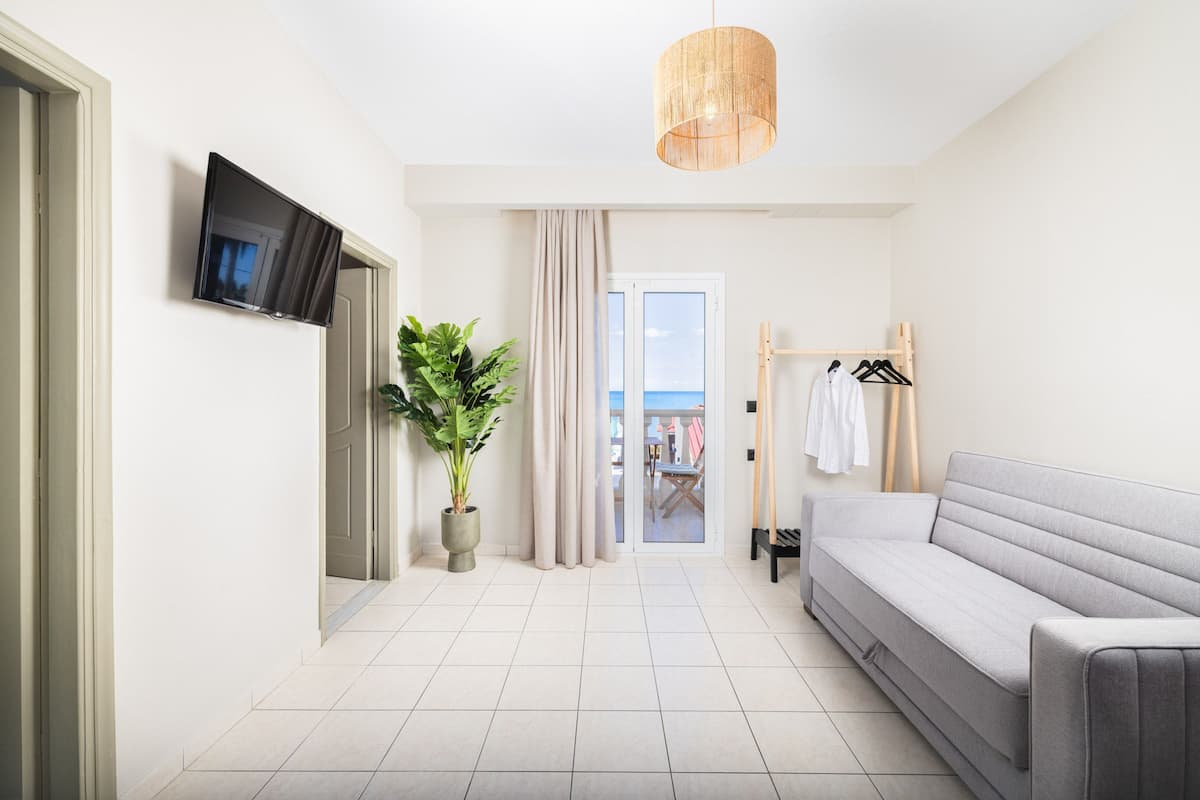 Una habitación de hotel moderna con una televisión de pantalla plana, una planta grande en maceta, un sofá gris, un perchero de madera y vistas al mar a través de un ventanal.