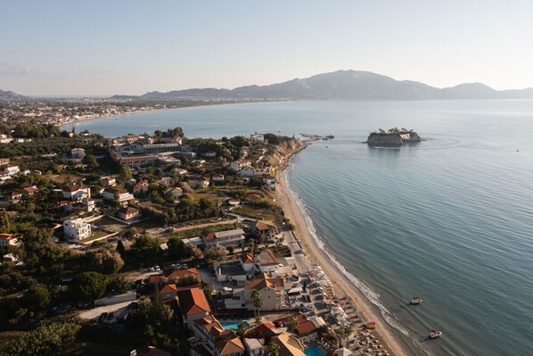 An aerial view of a coastal town with a beach, houses, and a small island. - Manthos Beachfront Apartments (Laganas)