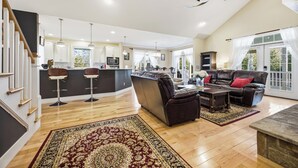 A spacious living area with a dark leather sofa, a wooden coffee table, and a patterned rug. A kitchen area with modern appliances and bar stools is visible in the background.