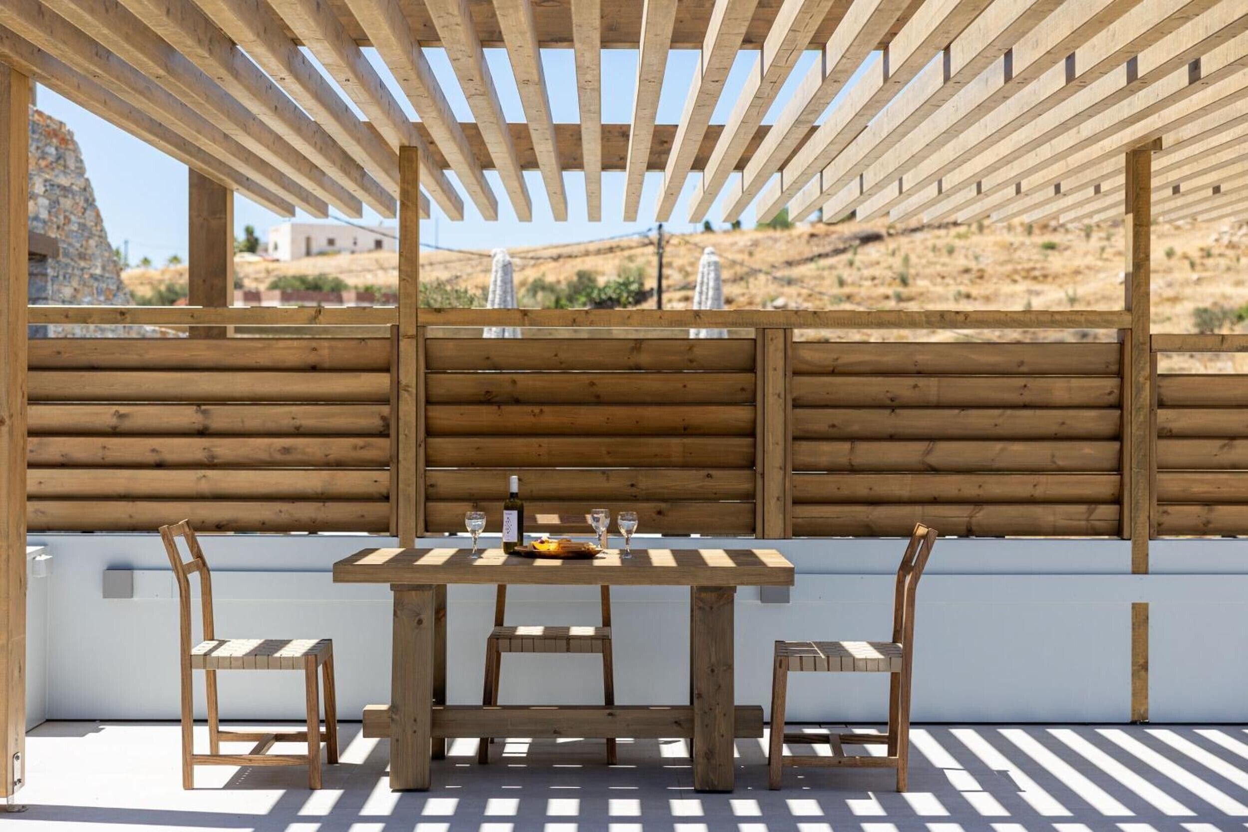 Eine hölzerne Pergola im Freien mit Tisch, Stühlen, einer Weinflasche und einem Teller Essen.
