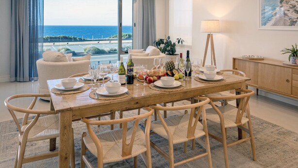 A dining area with a wooden table set for six, surrounded by wicker chairs. A large window offers a view of the ocean and a road. - Oceans Edge by Wine Coast Holidays (ALDINGA BEACH)