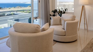 A modern living room with a large window offering a view of the sea and a road. There is a beige sofa and a floor lamp. - Oceans Edge by Wine Coast Holidays (ALDINGA BEACH)